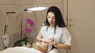 Genç ve güzel bir kozmetik uzmanı, bir güzellik salonundaki masaj masasına gözlerini kapatarak uzanan bir müşterinin tıbbi köpük ve masaj hareketleriyle yüzünü temizler.