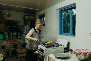 Yaşlı, beyaz, güzel bir kadın bir yaz günü eski bir kır evinin sobasında, patates hasadıyla patates kızartma tavasında spatulayla karıştırarak patates kızartıyor.