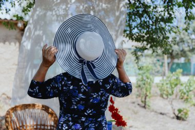Mavi elbiseli güzel, beyaz, yaşlı bir kadının portresi, ellerini tutan bir hasır şapka ve temiz havada sırt üstü duran bir köyün bahçesindeki bir ağacın altındaki gazlı bez perdesinin yanında.