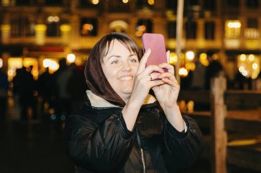 Bir bahar gecesi Brüksel 'deki Grand Place' de binaların mimarisinin fotoğraflarını çeken güzel, mutlu beyaz bir kızın portresi. Akıllı telefonuyla.