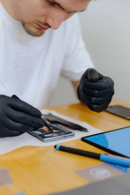 Beyaz tişörtlü ve siyah lateks eldivenli yakışıklı beyaz bir adam cımbızla akıllı bir telefona yuvarlak bir parça yerleştirir, bir yaz günü mutfakta bir masada oturur,