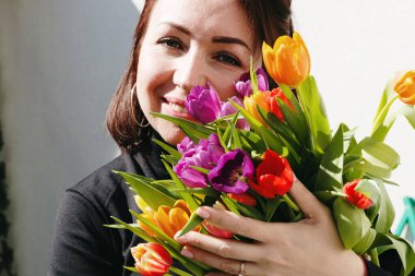 Kameraya gülümseyen ve yüzüne bir buket çok renkli lale bastıran güzel, mutlu, beyaz bir esmer kızın portresi. Bir bahar günü, beyaz bir duvarın yanında duruyor.