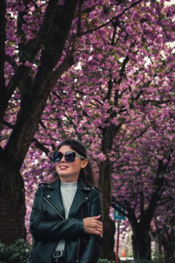 Güneş gözlüklü güzel beyaz bir kızın portresi ve gülümseyen deri ceketli bir şehir caddesinde çiçek açan sakura ağaçlarının arka planında duruyor..