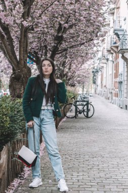 Beyaz saçlı, yeşil ceketli ve açık renk kotlu güzel bir esmer kızın portresi. Elinde çiçekli bir sakurayla şehir caddesinde dikilirken saçlarını düzeltiyor.