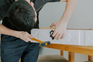 Koyu renk elbiseli esmer beyaz bir adam, bir yaz günü mutfakta, yakın çekimde, siyah saplı antika bir çiviyi, sarı bir çekiçle beyaz bir çekmeceye çakıyor. Kavram