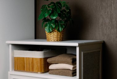 Doğal keçi kılından yapılmış çıplak gözlüklü ince süveterler, eski beyaz bir dolabın rafında, bir bambu sepetinin yanında katlanmış bir vazo içinde, kahverengi bir duvara yaslanmış bir çiçek, orta boy.