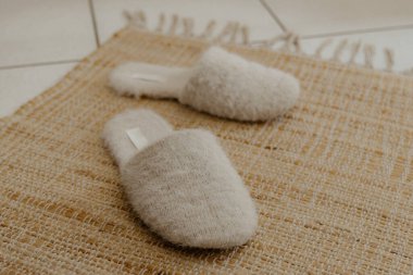 Bir çift güzel açık renkli bej renkli, yumuşak, modaya uygun ve sıcak terlikler odanın fayanslı zemininde doğal bir jüt halıda duruyor. Üst tarafı tarlanın derinliği ile yakın plan. Ev ayakkabıları