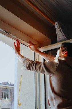 Genç, yakışıklı, esmer, beyaz bir adamın portresi. İki eliyle iç pencere çerçevesine özel kauçuk yapıştırıyor. Kış boyunca çatlakları kapatmak için.