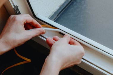 Pencerenin sol alt köşesine beyaz lastik izolasyon yapıştıran genç bir beyaz adamın portresi ikinci kattaki bir odanın siyah pervazının yanında, yakın plan yan manzara.