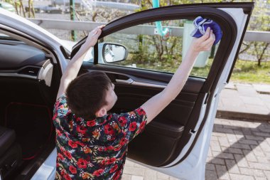 Güneşli bir bahar günü, araba yıkama yerinde, arka tarafta oturan ve arabanın kapısının içinden camı mavi bir bezle yıkayan beyaz bir adamın portresi. Konsept zaman aracı
