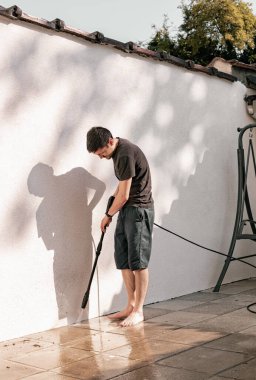 Siyah elbiseli esmer beyaz bir adamın portresi eğimli bir pozisyonda yalın ayak bahçe fayanslarını basınç altında su püskürterek temizlerken arka bahçedeki beyaz bir duvarın yanında bir kerpetenle