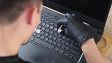 Siyah lateks eldivenli genç bir beyaz adam, bir yaz günü yatak odasında bir ofis masasında otururken dizüstü bilgisayarının klavyesine şeffaf bir harf etiketi yapıştırıp özel bir plastik fırça kullanıyor.