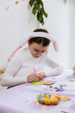 Tavşan bantlı güzel bir beyaz kızın göğsüne portre, masada oturmuş, kendi elleriyle bir kart yapıyor, paskalya yumurtalarına yapay elmasları yapıştırıyor ve bir bahar günü evde mutfakta bir civciv yapıştırıyor.