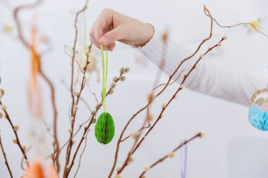 Portrait of one hand unrecognizable Caucasian girl hanging homemade green lace Easter paper egg on pussy willow branches, creating Easter bouquet on spring day, side view very close up selective focus. Concept of happy childhood, happy Easter
