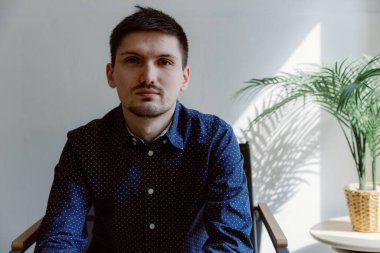 Mavi benekli tişörtlü, beyaz bir duvar ve bir saksıda bir palmiye çiçeğine karşı ofiste oturan, beyaz benekli, esmer, genç bir iş adamının portresi..