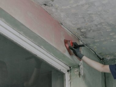 Eldivenli tanınmayan genç bir adam pencerenin yakınındaki kirli bir duvara fırçayla kırmızı çimento sürüyor. İskele üzerinde duruyor. Ev yenileme, inşaat işi kavramı.