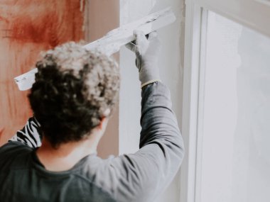 Üniformalı ve inşaat eldivenli beyaz saçlı genç bir adam pencere aralığını sıvamış, yakından bakıyor. Daire yenileme konsepti.