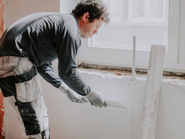 Gri tişörtlü ve kıvırcık kahverengi saçlı beyaz bir adam, pencerenin altındaki duvarda geniş bir spatula ile yeni macunu uyguluyor. Daire ve ev tadilatı, duvar sıva, sıva hizmetleri