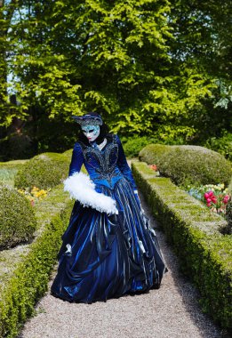 Grand Bigard Şatosu, Belçika - 27 Nisan 2025 Renkli koyu mavi Venedik kostümlü bir kadının tam boy portresi öğleden sonra Maya çiçek festivaline adanmış sergi sırasında parkta tek başına yürürken dinleniyor.