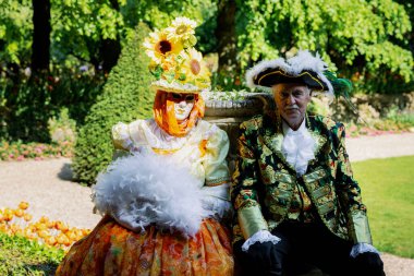 Grand Bigard Şatosu, Belçika - 27 Nisan 2025 Bir çiftin portresi, turuncu elbiseli, şapkalı kadın ve Venedik kostümlü adam, kameraya bakıyor ve sergi boyunca parkta birlikte dinleniyorlar.