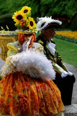 Grand Bigard Şatosu, Belçika - 27 Nisan 2025. Bir çiftin tam boy portresi, turuncu elbiseli kadın şapkalı ve Venedik kostümlü erkek Maya 'lara adanmış gündüz sergisi sırasında parkta tek başına oturuyor ve dinleniyor.