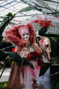 Grand Bigard Şatosu, Belçika - 27 Nisan 2025. Maya çiçek festivaline adanmış bir sergi sırasında, açık hava şemsiyesi ve tüylü pembe tonlu Venedik kostümlü bir kadının portresi bitkilerin arasında duruyor.