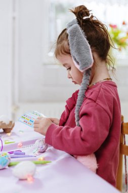 Paskalya yumurtalarına yanlamasına yapışkan yapıştırmalar yapıştıran, bahar günü mutfakta açık leylak renkli kağıt masa örtüsüyle masada oturan, gri tavşan bantlı güzel bir kız.