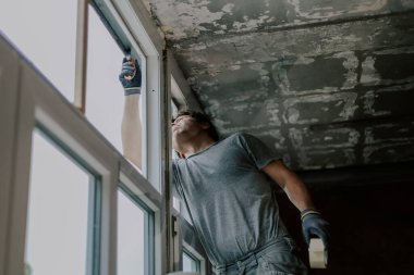Gri üniformalı yakışıklı beyaz bir adam, ikinci kattaki bahar bulutlu bir günde elinde bant tutarak ve inşaat bıçağıyla enkazı temizleyerek bir iskelenin önünde plastik pencere çerçevesini tamir ediyor.