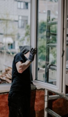 Siyah inşaat üniforması giyen yakışıklı beyaz bir adam yan yana duruyor, bir evin penceresini tamir ediyor ve kuruyor, yakından bakıyor. Ev yenileme ve pencere kurulumu kavramı.