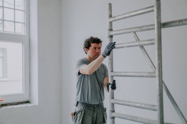 Genç, yakışıklı, beyaz, kıvırcık bir adam, tamirat ve inşaat çalışmalarının yapıldığı odaya bağımsız olarak metal iskeleler kuruyor. Ev yenileme ve pencere kurulumu kavramı.