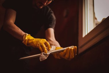Sarı eldivenli yakışıklı beyaz adam, plastik pencere çerçevesinin dar bir kısmını kuru bezle kurutuyor, pencerenin yanındaki evde dikiliyor, alt tarafı seçici bir odak ile yakın plan çekiyor. Ev yenileme ve pencere çerçevesi temizleme kavramı.