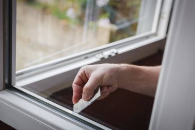 Bir elinde gri tişörtlü, tanınmayan beyaz bir adam çamurdan plastik pencere çerçevelerini keskinleştirici bir bıçakla temizliyor, bir bahar günü odanın içinde dikiliyor, yan görüntüsü çok yakın. Pencere temizliği, pencere çerçevesi tamiri