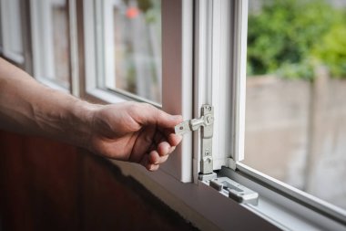 Bir elinde gri tişörtlü beyaz bir adam ayarlama yapmak ve plastik pencere çerçevesine kilitlenmek için metal bağlayıcı takıyor. Bahar günü odada duruyor, yan görüş çok yakın. Pencere temizleme, pencere çerçevesi tamiri, inşaat işi.