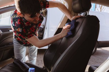 Güneş gözlüklü, beyaz tenli genç bir adamın portresi, eğilip arabanın ön yolcu koltuğunu güneşli bir bahar gününde araba yıkamada mavi bir bez parçasıyla temizliyor. Konsept araba yıkama, çalışma