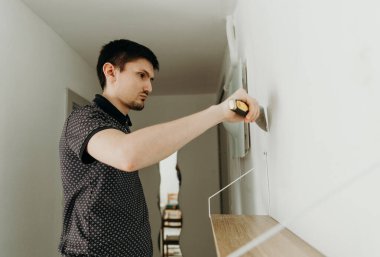 Beyaz tenli yakışıklı, esmer bir adam yarı yolda duruyor ve beyaz bir duvardaki vida deliğini spatula kullanarak kaplıyor, bir bahar günü bir odada duruyor, tarlanın derinliğine yakın bir açıdan bakıyor..