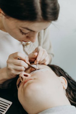 Genç ve güzel bir beyaz kadın kozmetik uzmanının portresi. Yapay kirpikler yapıştırıyor. Kadın bir müşterinin sol gözüne iki cımbız yapıştırıyor. Kirpik uzantısı