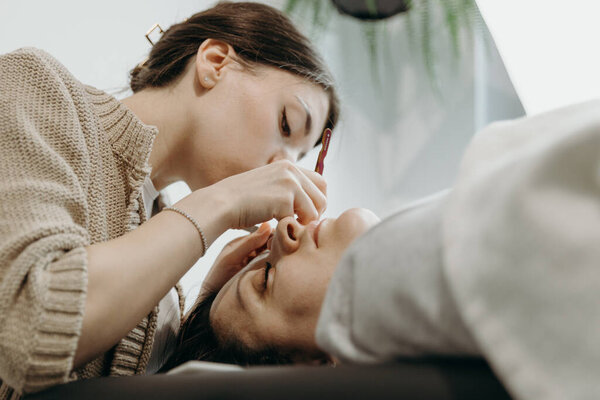 Портрет одной молодой белой брюнетки-косметолога, которая клеит искусственные ресницы двумя пинцетом на левый глаз клиентки, лежащей на косметологическом столе в домашнем офисе в весенний день, вид снизу. Реснички