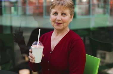 Masada yorgun bir şekilde oturan, elinde bir bardak meşrubat tutan ve alışveriş merkezinde alışveriş yaptıktan sonra dinlenen kameraya bakan, kırmızı bluzlu, beyaz, yaşlı ve güzel bir kadının portresi. Kavram, alışveriş zamanı, içki zamanı.