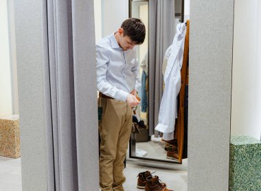 Beyaz tenli, bej pantolon ve açık mavi gömlekli genç, yakışıklı bir beyaz adam aynaya yansıyan bir soyunma odasında kemer takıyor. Çevrimdışı alışveriş kavramı, yaşam tarzı adamı.