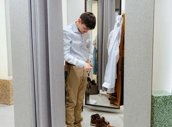 Beyaz tenli, bej pantolon ve açık mavi gömlekli genç, yakışıklı bir beyaz adam aynaya yansıyan bir soyunma odasında kemer takıyor. Çevrimdışı alışveriş kavramı, yaşam tarzı adamı.