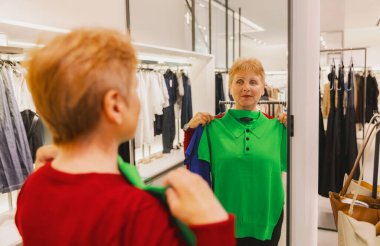 Güzel, beyaz, mutlu, kırmızı kazaklı yaşlı bir kadının portresi. Yeni yeşil tişört deniyor. Bahar günü dükkânda aynada kendine bakıyor. Zaman alışverişi, emeklilik yaşam tarzı, mutlu yaşlılar..