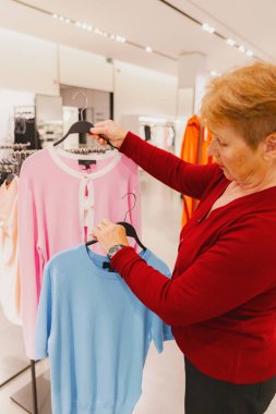 Beyaz, kırmızı kazaklı, güzel bir kadının portresi. Mavi tişört ve pembe hırka arasında seçim yapıyor. Bahar günü dükkânda duruyor. Konsept zaman alışverişi, emeklilik yaşam tarzı, kıyafet seçimi.