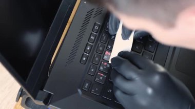 Siyah lateks eldivenli genç bir beyaz adam dizüstü bilgisayarın klavyesindeki şeffaf bir çıkartmayı temizlerken özel bir kağıt şeridi kullanarak bir yaz günü yatak odasında oturuyordu. Üst tarafı yavaş çekimde çok yakındı.