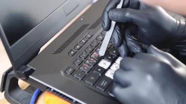 Siyah lateks eldivenli genç bir beyaz adamın elleri, dizüstü bilgisayarın tuşları üzerinde yürürken cımbızla şeffaf bir çıkartma yapıştırıyor, yaz günü yatak odasında bir ofis masasında oturuyor, yan görünümü çok yakın.