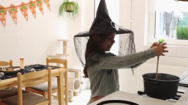 Cadıların şapkası ve siyah tüllü güzel beyaz bir kız, Cadılar Bayramı pastasını coşkuyla karıştırıp gaz sobasında, ahşap bir kaşıkla, bir tatil ziyafeti hazırlıyor, bir sonbahar günü mutfakta duruyor,