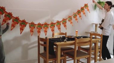 Kollarında kedi yavrusu olan üç güzel beyaz kız, beyaz bir mutfak duvarını şenlikli bir dekorla süslüyor - turuncu Cadılar Bayramı bayrakları, onları sonbahar günü çift taraflı bantlara yapıştırıyor, yavaş çekimde. Evi dekore etme zamanı.