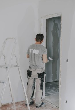 Gri üniformalı yakışıklı beyaz bir adam ve arkadan gelen eldivenler kapının önünde dikilirken elinde küçük bir spatula ve macun tutuyor. İnşaat konsepti.