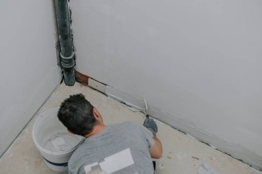 Gri üniformalı genç bir beyaz adam merdivenlerde diz çöküp duvarın altını küçük spatulayla kaplıyor. Üst tarafı seçici bir odak noktasıyla kaplıyor. Sıva ve inşaat işi kavramı.