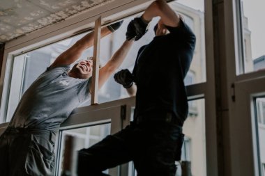 Üniformalı iki yakışıklı beyaz adam bir odada iskele üzerinde duruyor ve birbirlerine duygusal olarak yardım ediyorlar, plastik çerçevelerdeki çatlakları onarmak için bahar akşamları İskoç bandı kullanıyorlar, yakın plan çekimde. Pencere onarımı ve kurulumu