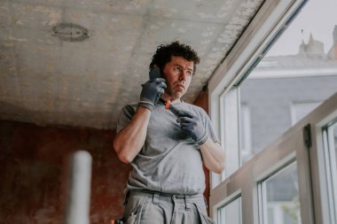 Gri üniformalı yakışıklı beyaz bir adam, yenileme çalışmalarının devam ettiği bir evin penceresinin yanındaki iskelenin yanında duruyor ve duraklama sırasında cep telefonuyla dikkatlice konuşuyor. Ev yenileme konsepti, pencere